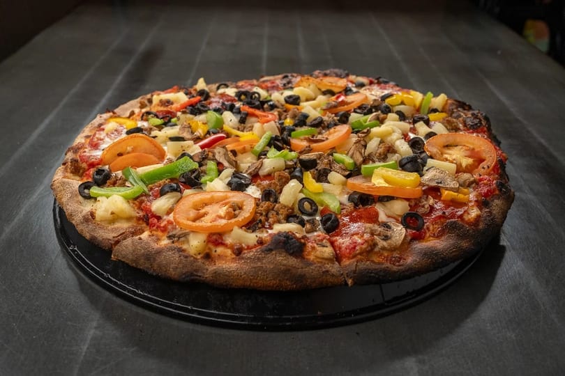 A whole pizza with various toppings including tomatoes, olives, peppers, and pineapple on a dark surface.